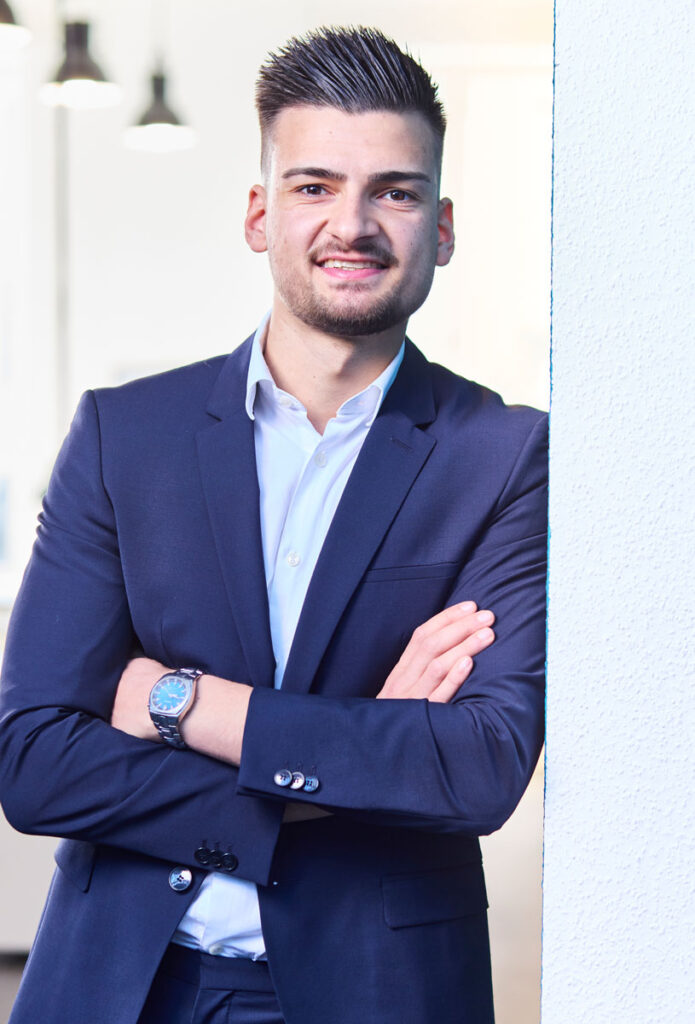 Ein lächelnder Mann im blauen Anzug lehnt an einer Wand, trägt eine Armbanduhr.